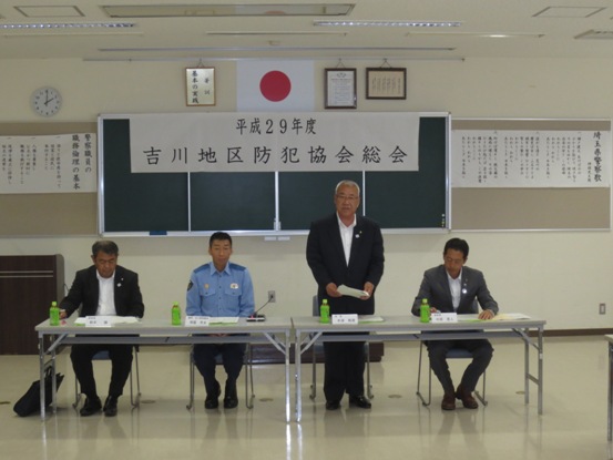「平成29年度吉川地区防犯協会総会」と書かれているポスターが貼りつけられた黒板の前で、吉川地区防犯協会会長としてあいさつの言葉を述べる三郷市長と、席札とペットボトルのお茶が置かれた市長と横並びの席に座り資料に目を通しながら話を聞く関係者3名の写真