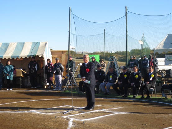 晴天の下、グラウンドに立ち開会の挨拶の言葉を述べる三郷市長と、後ろのグリーンネットの近くに置かれたパイプ椅子に座り話を聞く黄色い胸章を付けた関係者の写真