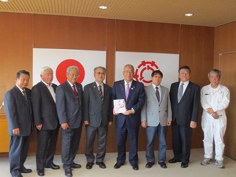 国旗と三郷市旗が飾られた室内で、目録を持った市長と7名の関係者が横一列に並んでいる記念写真