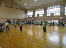 体育館の壁や窓側の内周に参加者が集まり、中央にゼッケンをつけた参加者が間隔をあけて立っている借り物競争の様子の写真