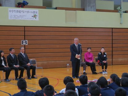 左右に3名ずつ椅子に座った関係者の間のマイクスタンドの前で挨拶を行っている市長の話を、向かい合って座っている生徒たちが聞いている後ろ姿の写真