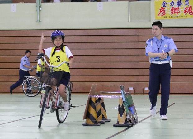 紫色のヘルメットを被り黄色のゼッケンをつけて自転車に乗っている女の子が障害物の左側で右腕を直角に上に伸ばしている様子を警察官がチェックしている実技の様子の写真