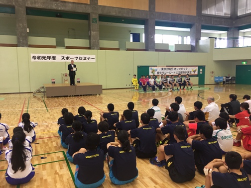 「令和元年度 スポーツセミナー」と書かれた横断幕が壁に貼られた体育館で、可動式の小さなステージの上に乗り挨拶を行っている市長の話を、向かい合い体育座りをして聞いているユニホームを着た子供たちの写真