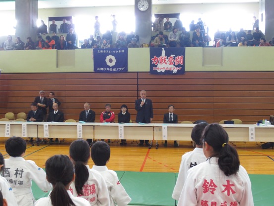 紺地に白い花のマークと紺地に赤字で「力戦奮闘」と書かれている2種類の旗が、たくさんの観客がいる2階席から掛けてある壁を背に、長机が一列に並べられた関係者席を立ち挨拶の言葉を述べる三郷市長と、緑のマットの上に整列して立って話を聞くそれぞれの名前の入ったゼッケンが付いている柔道着を着用した子どもたちの写真
