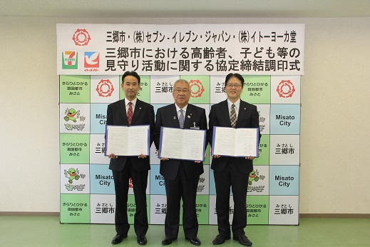 「三郷市における高齢者、子ども等の見守り活動に関する協定締結調印式」と書かれたポスターが貼ってある三郷市のバックボードの前で、三郷市長と株式会社セブンーイレブン・ジャパン、株式会社イトーヨーカ堂の社員が協定書を手に持ち記念撮影をしている写真