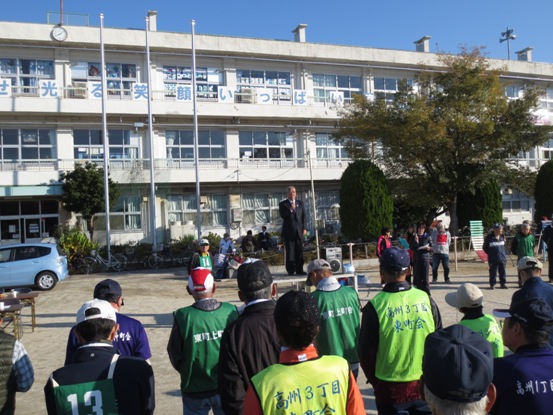 高州小学校の校庭に設置された朝礼台に立ち激励の言葉を述べる三郷市長と、話を聞く緑と黄緑のゼッケンと帽子を着用したグラウンドゴルフ大会参加者の写真