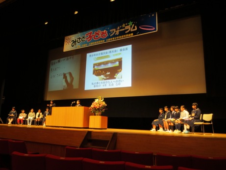「みさと子どもフォーラム」と書かれた横断幕の後ろに設置されたスクリーンに映像が映し出され、演壇に立っている子供が発表を行っている様子の写真