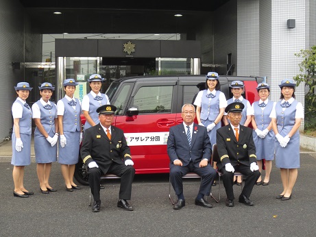 建物の前に駐車した赤色のボディの車の前に、薄水色の制服を着用したアザレア分団の方々が左右に4名ずつ立ち、手前に市長を中央に2名の制服姿の消防団員の男性が座っている記念写真