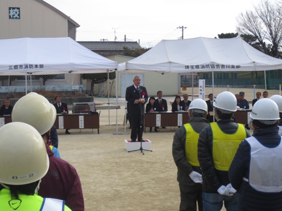 白のテントが張られた校庭で、ヘルメットを被った関係者たちが整列しているなか、正面に立つ市長が挨拶をしている写真
