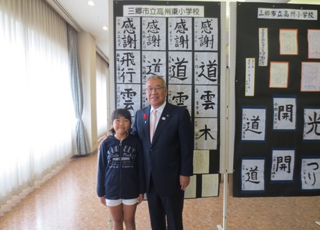 学校別に掲示ボードに展示されている書道の作品の前で、紺色のパーカーを着た女の子と市長が横に並んで立っている記念写真