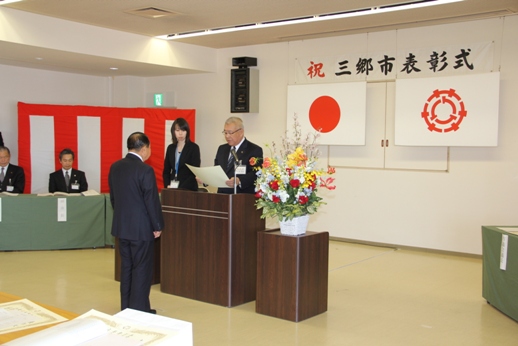 「祝 三郷市表彰式」と書かれた会場で演壇を挟んで立っている男性に向けて市長が賞状を読み上げている写真