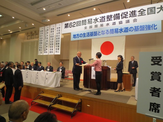 「第62回簡易水道整備促進全国大会」と書かれたボードの下に「地方の生活基盤となる簡易水道の基盤強化」と書かれたボードと国旗が吊るしてあるステージの演台の前で、ステージの左前方に設けられた来賓席を背に、女性から表彰状を受け取る三郷市長の写真