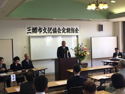 「三郷市文化協会定期総会」と書かれたポスターがホワイトボードの上に貼ってあり、オレンジ・黄色・紫・緑の花々で彩られた花台と並ぶ演台に立ち話をする三郷市長と話を聞く司会進行役と参加者の写真