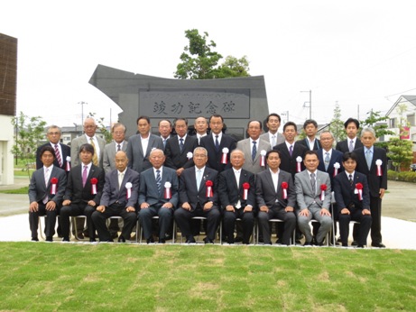 竣功記念碑の前に設置された椅子に市長を中央に9名の関係者が座り、後列に15名の関係者が立っている記念写真