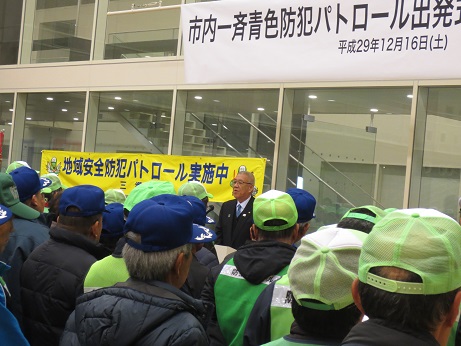 「市内一斉青色防犯パトロール出発式」と書かれたポスターとかいちゃん・つぶちゃんのイラストとともに「地域安全防犯パトロール実施中」と書かれた横断幕が貼り付けられたガラス壁の前で、挨拶の言葉を述べる三郷市長と、向かい合って立ち話を聞く緑と紺のキャップを着用した町会等自主防犯団体のメンバーの写真