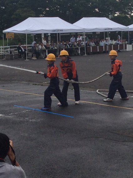 オレンジ色の作業着にヘルメットを被った3名の少年消防クラブ員が力を合わせてポンプ操法を行っている様子の写真
