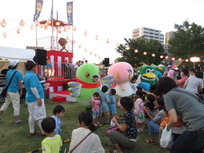 公園の中央に設置された櫓の前でマスコットキャラクターかいちゃんやピンク色や緑色の近隣のキャラクターが並び、小さな子供たちが一緒に母親から撮影してもらっている様子の写真