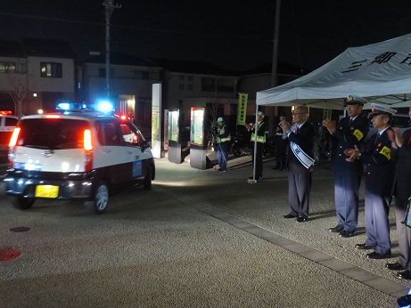 暗闇の中、白いバザーテントの前に立ち、青い回転灯を付けたパトロールカーを見送るタスキをかけた三郷市長と警察官の写真