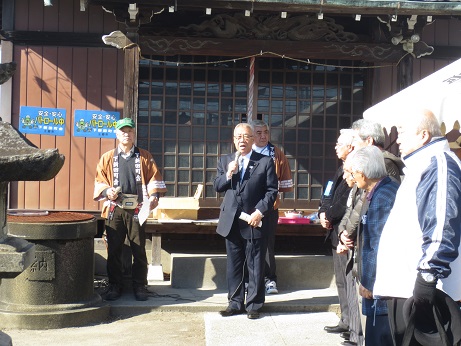 下新田公民館神社前広場で、マイクを手に持ち挨拶の言葉を述べる三郷市長と「下新田町会」の文字が入った法被を着用し市長の後ろに立つ男性2名、白いバザーテントの前に立ち市長の話を聞く参加者の写真