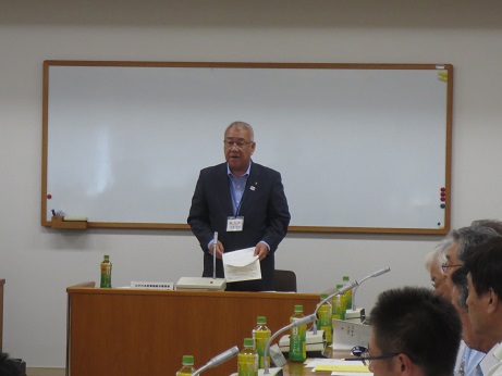 ホワイトボードの前に配置された演台に立ちあいさつの言葉を述べる三郷市長と、ペットボトルのお茶とマイクが置かれた席に座り話を聞く参加者の写真