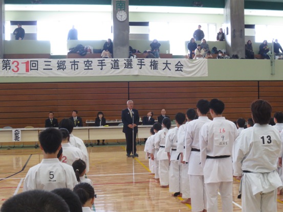 「第31回三郷市空手道選手権大会」の横断幕が壁に貼られている体育館で、道着を着用した参加者たちが整列しているなか、正面のマイク台の前に立って挨拶をする市長の写真