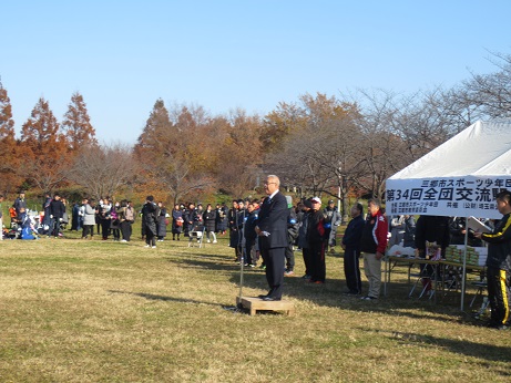 「三郷市スポーツ少年団第34回全団交流駅伝大会」と書かれた横断幕が白いバザーテントに貼り付けてあり、テント前に設置されたお立ち台に立ち挨拶の言葉を述べる三郷市長と周りに立って話を聞く関係者の写真