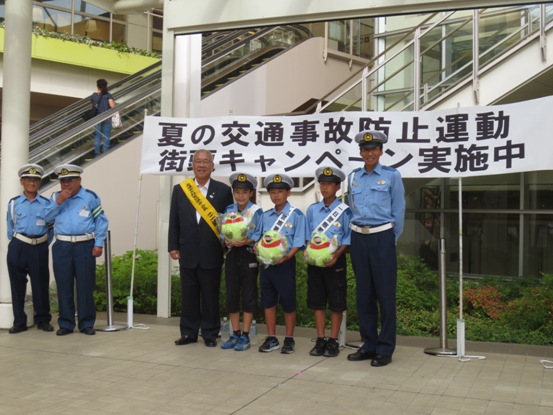「夏の交通事故防止運動街頭キャンペーン実施中」と書かれた横断幕の前で、黄色いタスキをかけた三郷市長がかいちゃんのぬいぐるみを両手に持った一日警察署長の子どもたちと警察官の男性とともに記念撮影をしている写真