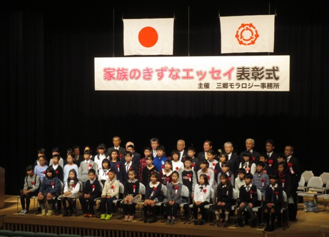「家族のきずなエッセイ表彰式」と書かれた横断幕と国旗・三郷市旗がステージ上に吊るしてあり、胸章を付けたたくさんの入選者の子どもたちと三郷市長・関係者が並んで記念撮影をしている写真