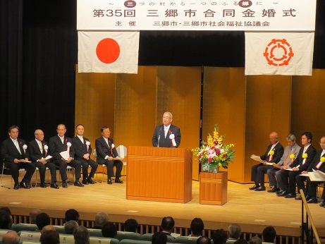 「第35回三郷市合同金婚式」と書かれた横断幕と国旗と三郷市旗が飾られ金屛風が設置されたステージの左右に胸章リボンをつけた関係者が座り、中央の演壇で挨拶を行っている市長の写真