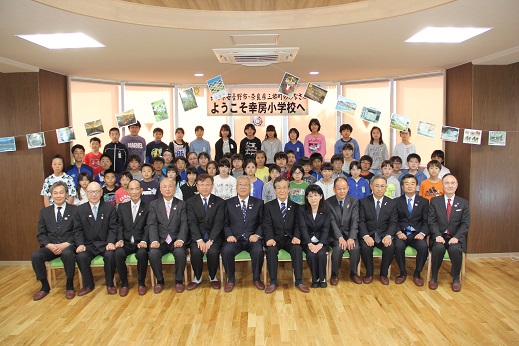 「ようこそ幸房小学校へ」と書かれたポスターとフォトガーランドが飾られた部屋で、前列に三郷市長と協議会メンバー、後列に幸房小学校の子供たちが並び記念撮影をしている写真