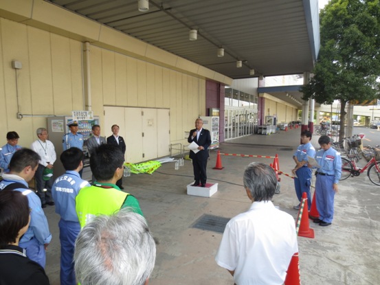 ピアラシティ横で、白い踏み台に立ち挨拶の言葉を述べる三郷市長と、話を聞く国道298号違法駐車・ポイ捨て対策検討会のメンバーの写真
