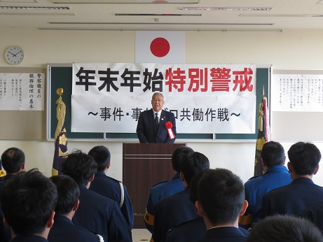 「年末年始特別警戒～事件・事故ゼロ共働作戦～」と書かれた横幕が貼られた室内で、警察官の方が並んで座っている前の演壇に立ち挨拶を行っている市長の写真