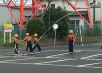 オレンジと紺の制服と黄色いヘルメットを着用した少年消防クラブのメンバーが、白線の引かれた駐車場で消防ホースをともに持ち放水している写真