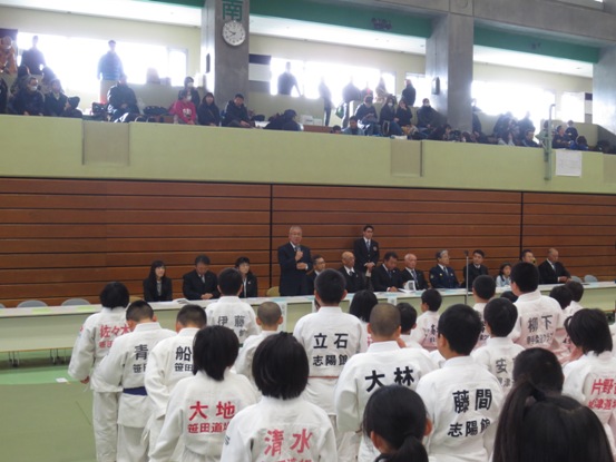 体育館の壁側に長机が並び、席から立ち上がり挨拶を行っている市長の話を、向かい合って立っている道着を着用した子供たちが聞いている後方から写した写真