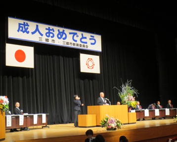 「成人おめでとう」と書かれた横断幕や国旗と三郷市旗が飾られたステージの左側に黒色のスーツを着た女性の手話と同時に檀上に立っている市長が式辞を述べている様子の写真