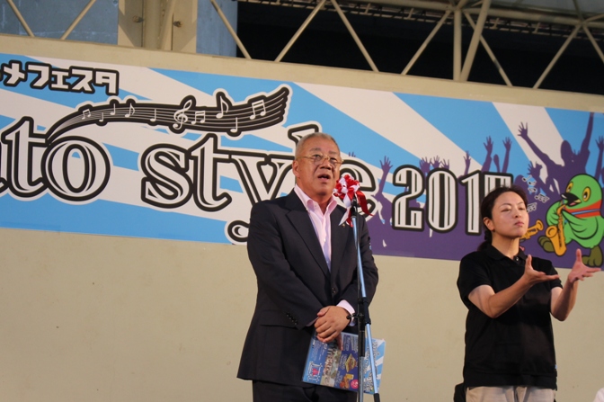 黒色のシャツを着た女性の手話と同時にマイクスタンドの前に立ち挨拶を行っている市長の写真