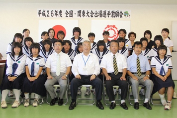 平成26年度全国・関東大会出場選手激励会と書かれた横断幕、国旗と三郷市旗が飾られた室内で女子ハンドボールで優秀な成績を収めた北中学校の3名の女子生徒と3名の男性が市長を挟んで横一列に並んで椅子に座り、後方に15名の生徒と5名の女性が立っている記念写真
