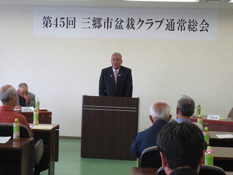 「第45回三郷市盆栽クラブ通常総会」と書かれたポスターが壁に貼ってあり、その前に設置された演台に立って話をする三郷市長と、ペットボトルのお茶が置かれた席に座り話を聞く参加者の写真