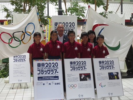 「東京2020オリンピック・パラリンピック フラッグツアー」と書かれた立て看板が3つ置かれ、その後ろに並ぶワインレッドのユニフォームを着用した子どもたちと三郷市長、関係者の女性がオリンピック・パラリンピックの大きな旗とともに記念撮影をしている写真