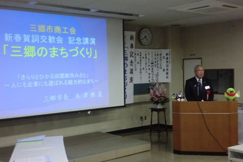 スクリーンに「三郷のまちづくり」と題した資料が映し出され、左側の演壇に立つ市長が講演を行っている様子の写真