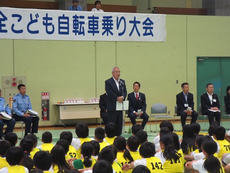 前に立ち開会のあいさつをする三郷市長と、話を聞く黄色いゼッケンを着用した小学生とパイプ椅子に座った関係者の写真