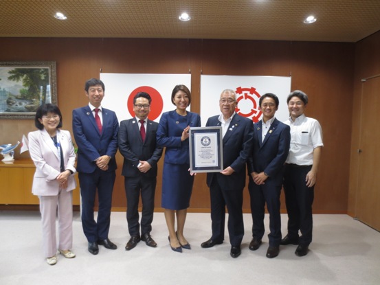国旗と三郷市旗が飾られた室内で、青色のスーツを着た女性と市長が額入りの認定証を持ち、5名の関係者と横一列に並んでいる記念写真