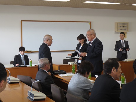 平成29年度第1回三郷市地域公共交通活性化協議会で、手に持った文書の内容を読み上げている三郷市長と立って聞く男性、三郷市長の横で黒いトレーに入った文書を確認している女性の写真