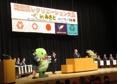 「埼玉県レクリエーション大会inみさと」と書かれた横断幕と国旗・三郷市旗を含む5つの旗がステージ上に吊るしてあり、赤・黄色・白の豪華な花々で彩られた花台と並ぶ演台に立ち挨拶の言葉を述べる三郷市長と、演台の横で並んで立っている、手話通訳をしている女性、かいちゃん、つぶちゃんを両手に抱いた女子学生、ステージ上に配置された席に座り話を聞く胸章を付けた来賓者たちの写真
