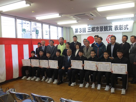 「第3回三郷市景観賞表彰式」と書かれた横断幕と国旗・三郷市旗が貼り付けられている壁の前で、前列に置かれたパイプ椅子に三郷市長を中心に座る表彰状を両手に持った8名の男子学生と、後ろに並んで立つ関係者たちが記念撮影をしている写真