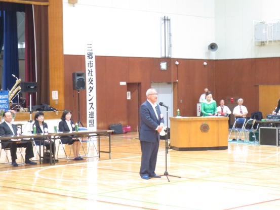 「三郷市社交ダンス連盟」と書かれた垂れ幕が勤労者体育館の壁に貼り付けてあり、セレモニーで挨拶の言葉を述べる三郷市長とペットボトルのお茶が置かれた席に座り話を聞く関係者の写真