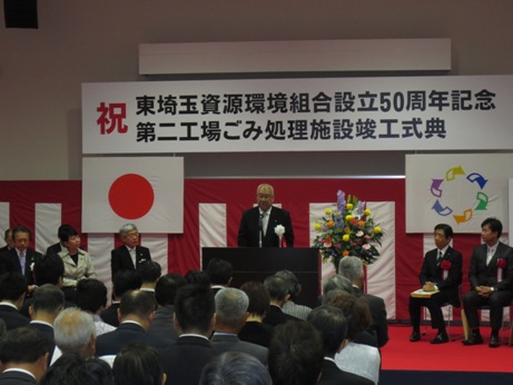 「祝 東埼玉資源環境組合設立50周年記念、第二工場ごみ処理施設竣工式典」と書かれた横断幕と紅白幕が貼られた会場の演壇で挨拶を行っている市長の話を聞いている、胸章リボンをつけた関係者や参加者の写真