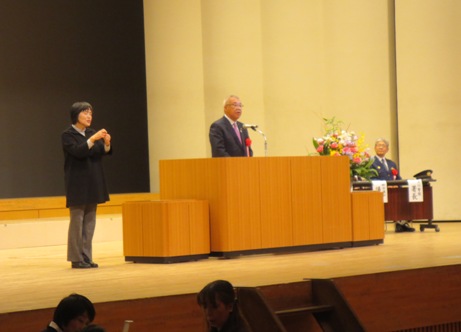 ステージの右側に設置された席に座っている関係者の方が見守るなか、演壇の左側に立つ手話通訳者の女性が手話を行いながら演壇に立っている市長が挨拶を行っている写真