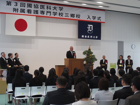 国旗と獨協医科大学の校旗の上に「第3回獨協医科大学附属看護専門学校三郷校入学式」と書かれた横断幕が掛けられ、盆栽が置かれた花台と並ぶ演台に立ちあいさつの言葉を述べる三郷市長と、座って話を聞く赤と白の胸章を付けた関係者と入学生の写真
