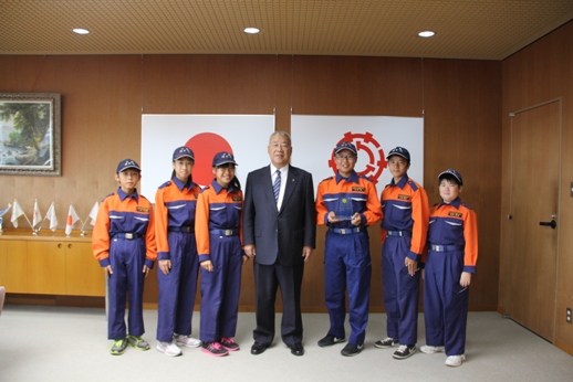 国旗と三郷市旗が飾られた室内で、市長を挟んで6名のクラブ員が立っている記念写真